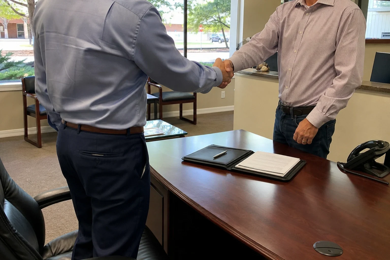 Two men in button-down shirts shake hands beside a wooden desk with an open padfolio and pen, waiting area chairs and windows behind in a small office